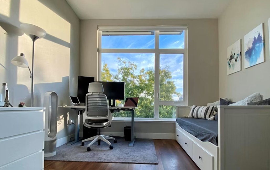chambre avec petit lit à droite et bureau avec chaise ergonomique et plusieurs écrans face à la fenêtre avec vue sur des arbres