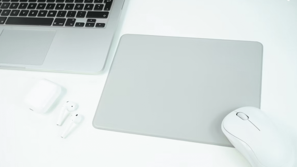 tapis de souris gris posé sur une table blanche avec une souris et des écouteurs sans file blancs à coté d'un pc portable